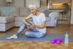 Une femme aînée lisant sur son téléphone, assise sur un tapis de yoga à côté de petis altères et d'une bouteille d'eau.
